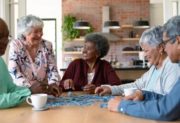 Group of five people smiling and working together to assemble a jigsaw puzzle on a brightly lit dining room table.