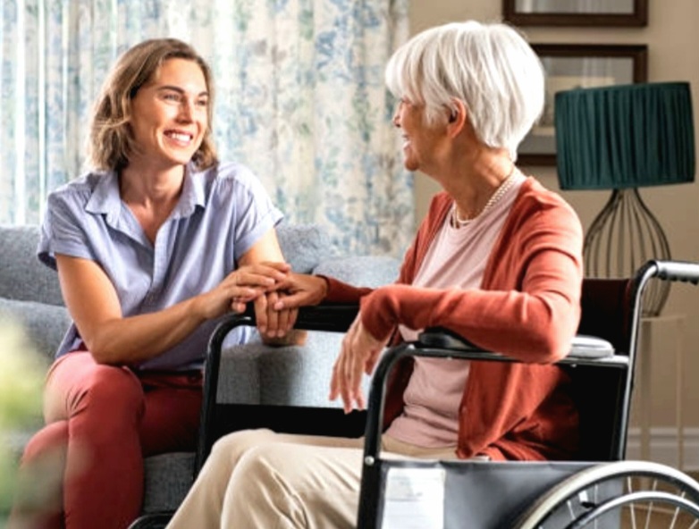 A compassionate female caregiver sitting on a couch, holding hands with an elderly woman in a wheelchair.